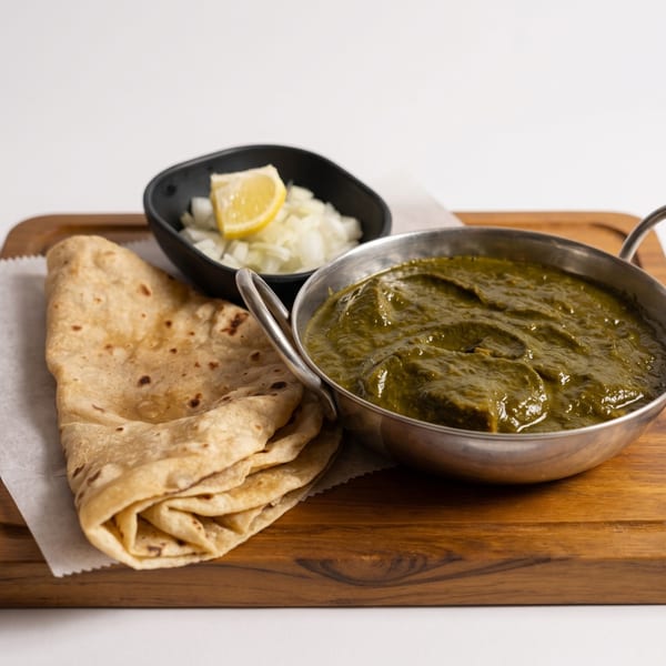 A metal bowl of green curry, flatbread, and a side of onions with lemon on a wooden board.