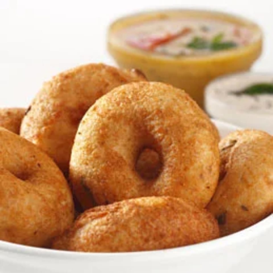 A bowl of golden-brown medu vada, with two small bowls of chutney and sambar in the background.